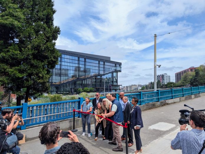 Harrell holds ceremonial gold scissors to cut the red ribbon on a widen sidewalk next to a new protected bike lane on Pike Street on the I-5 overpass bridge to Capitol Hill. The new convention center is in the background.