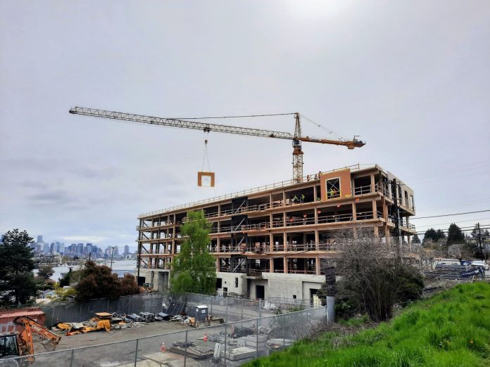 The mass timber framework is visible on a six-story office building as a tower crane lowers a siding panel into place.
