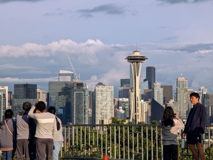 A group of young people stand by the railing at Kerry Point with the Space Needle and downtown skyscrapers looming in the distance.