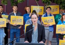 Wilson Defeats Harrell Wilson stands at a lectern speaking and a dozen supporters stand behind her holding yellow Wilson for Mayor signs on the City Hall Plaza.