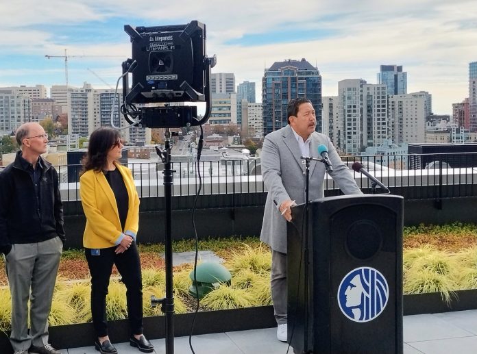 Harrell stands at a lectern with the Seattle City Seal and the skyline visible behind him from a midrise rooftop.