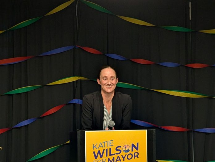 Wilson stands in front of a lectern with her yellow campaign sign with rainbow streamers behind her.
