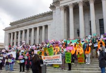 Op-Ed: Tech Workers Must Challenge the Political Power of Their Bosses More than 100 advocates hold signs urging a balanced tax code on the steps of the state Capitol building with its many marble columns.