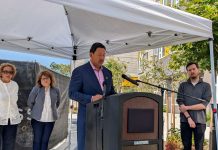 Harrell Announces Reparations Fund, Ramps Up Attacks on Wilson Harrell stands at a lectern under a white tent with Bryant Manor apartment building in the background.