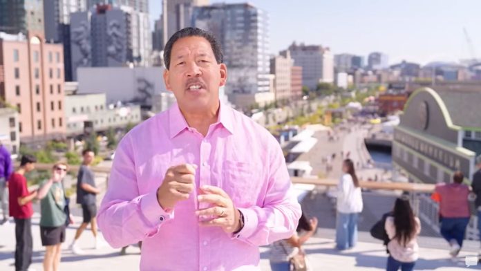 Mayor Harrell 2025 speech on budget proposal via Seattle Channell Harrell weather a pink button up shirt with pedestrians on the Overlook Walk in the background.