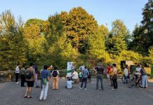 Kicking off The Urbanist Fall Subscriber Drive, Score Your Merch A group of about 30 people stands in a park with an Urbanist banner set up and a stand of trees in the background.