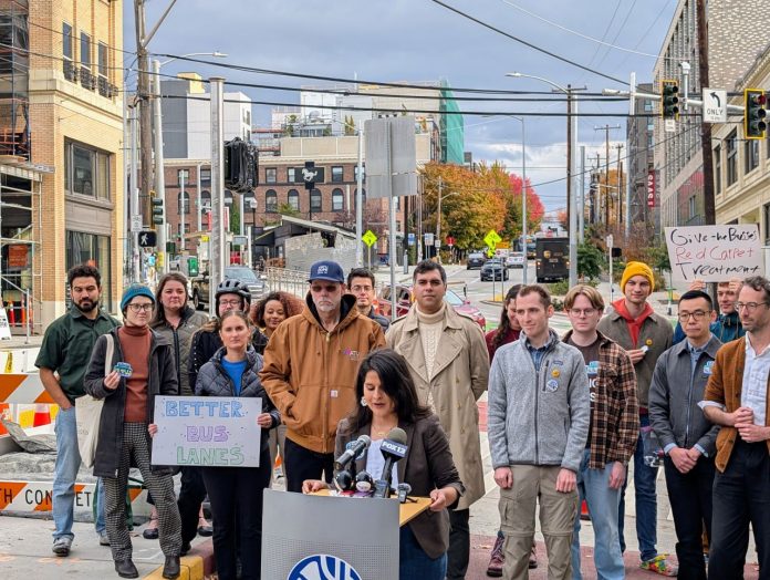 More than two dozen advocates stand behind Rinck at the bus stop at 12th Avenue and Union Street, with Madison Street in the background.
