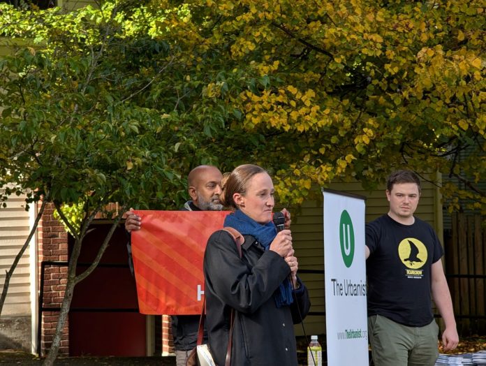Wilson holds a mic and two volunteers who banners for The Urbanist and for Seattle Tech4Housing.