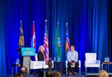 Op-Ed: Ferguson Must Back ‘Stand Up to Trump’ Rhetoric with Funding Ferguson and Smith sit on white lounge chairs with a row of flags behind them for the Cascadia states and nations.