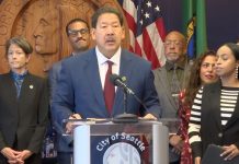 Harrell Vows to Block ICE Overreach, but Plan Lacks Teeth Harrell wears a suit and stands at a lectern with the seal of Seattle. He's surrounded by city councilmembers and staffers including Sara Nelson, Rob Saka, and Mark Solomon.