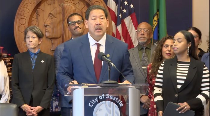 Bruce Harrell ICE press conference - Seattle Channel Harrell wears a suit and stands at a lectern with the seal of Seattle. He's surrounded by city councilmembers and staffers including Sara Nelson, Rob Saka, and Mark Solomon.