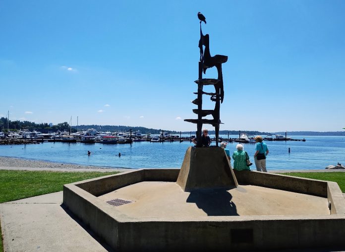 Kirkland waterfront A tower-like metal sculpture crowns Kirkland's Marina Park, with a few pedestrians walking on the path along its edge. The marina and Moss Bay is in the distance.