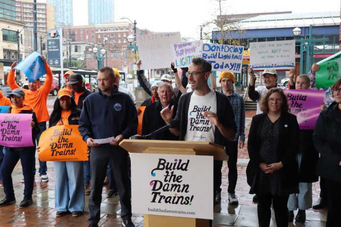 Advocates wave "build the damn trains sign" and Kirk from Transportation Choices Coalition speaks at the lectern