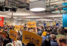 Wilson Trails in Early Results, Seattle Mayor’s Race on Nailbiter Course The view from the back of the room with supporters waving yellow Wilson signs and the candidate in the distance on stage.