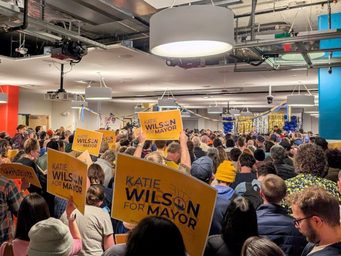 The view from the back of the room with supporters waving yellow Wilson signs and the candidate in the distance on stage.