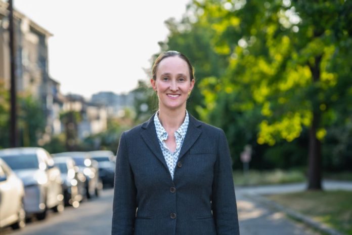 Katie Wilson for Seattle campaign photo Wilson wears a blzer and stands on an urban street with trees and a line of parked cars in the background.