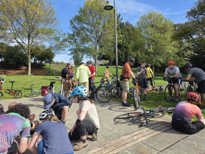 About a dozen people are pictured sitting or standing with their bikes in a park.