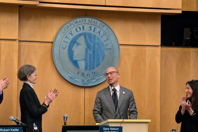 Lin wears a gray suit and glasses and stands at the lectern with the City of Seattle seal behind him. Council President Sara Nelson and Maritza Rivera are to either side clapping.