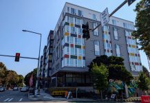 Seattle Leads Nation in Affordable Apartment Production A seven story apartment building with gray and white siding with red, gold, and green accents. Photo shows the view from the nearby intersection.