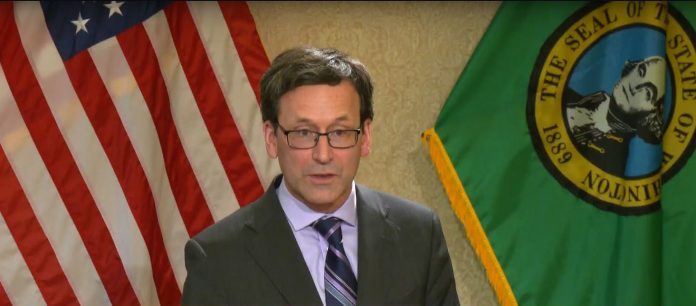 Ferguson wears a dark gray suit and tie and stands in front of the flags of the United State and Washington state.